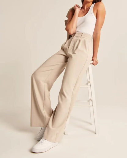 High-waist pleated wide-leg pants in beige, styled seated on a stool